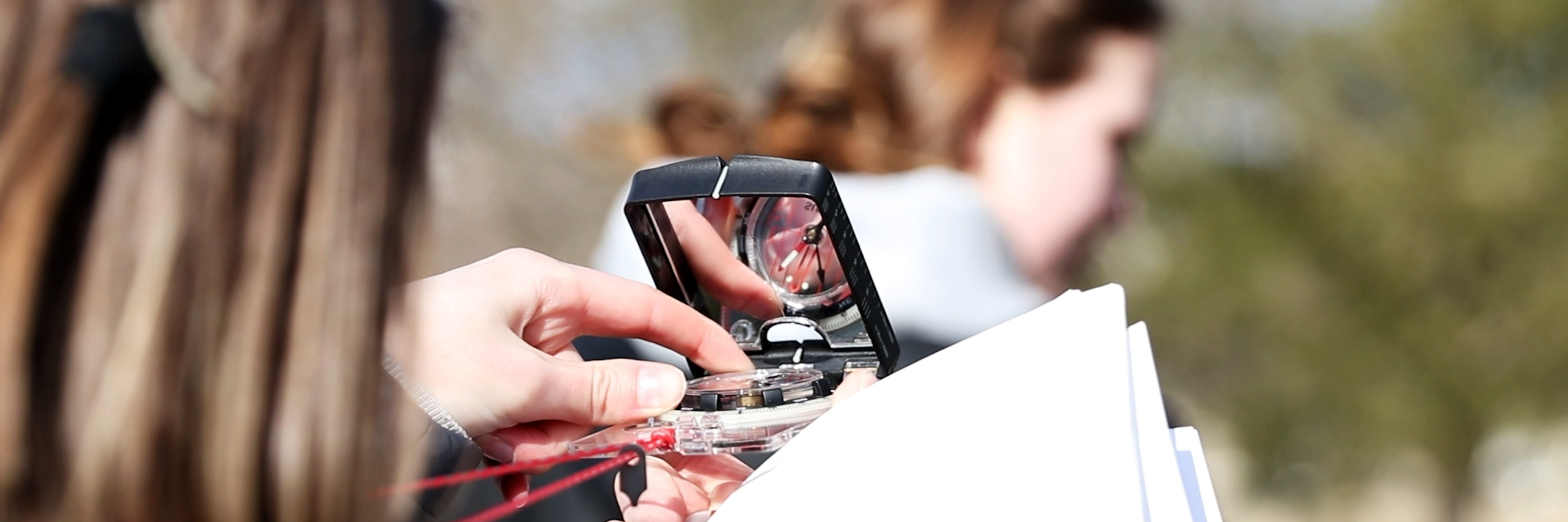 A closeup of a student's hands in a forest holding a compass to eye level while turning the dial